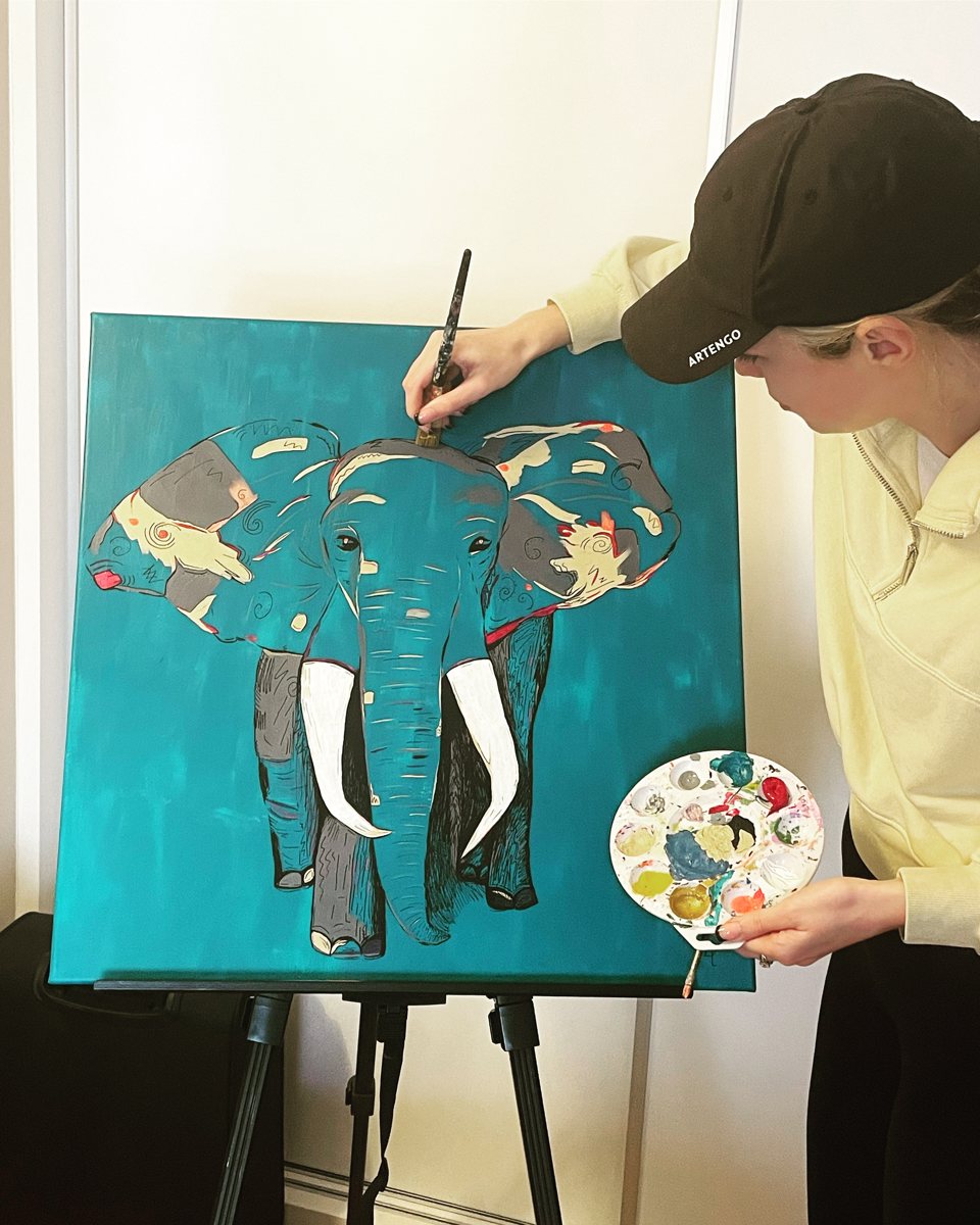 Niamh at her easel, brush in hand, painting the teal elephant with a palette of paint in her other hand